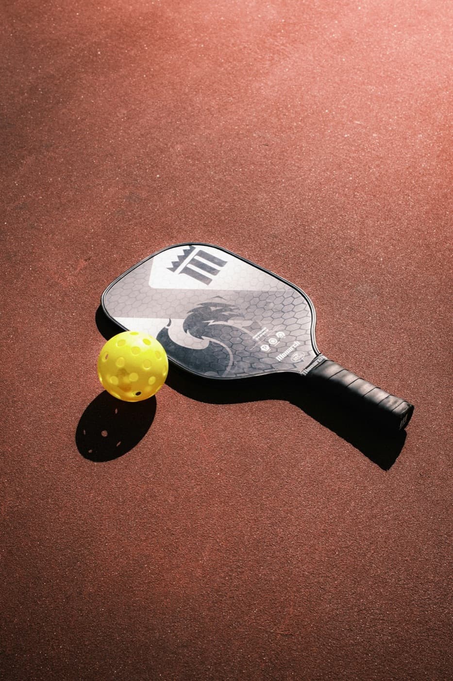 A pickleball paddle and a yellow ball on a clay coloured court.